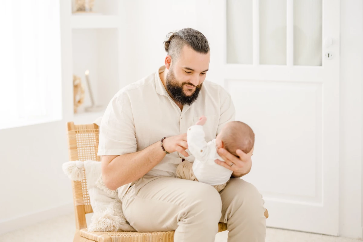 Séance nouveau-né en studio : jeune papa nourrit son bébé au biberon dans une ambiance lumineuse et épurée, moment de tendresse paternelle