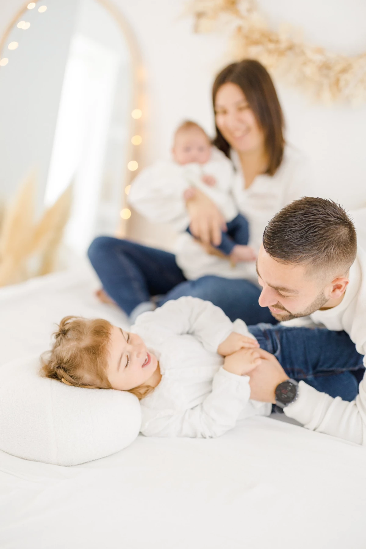 Séance naissance en studio : famille complice avec nouveau-né, papa joue avec son enfant dans une ambiance douce et lumineuse