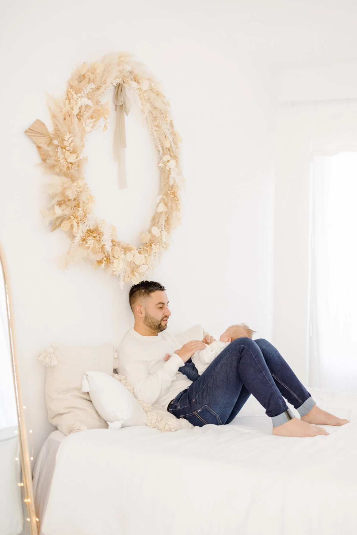 Séance naissance en studio : jeune papa assis sur le lit avec son bébé, ambiance épurée et lumineuse en tons blancs et beige