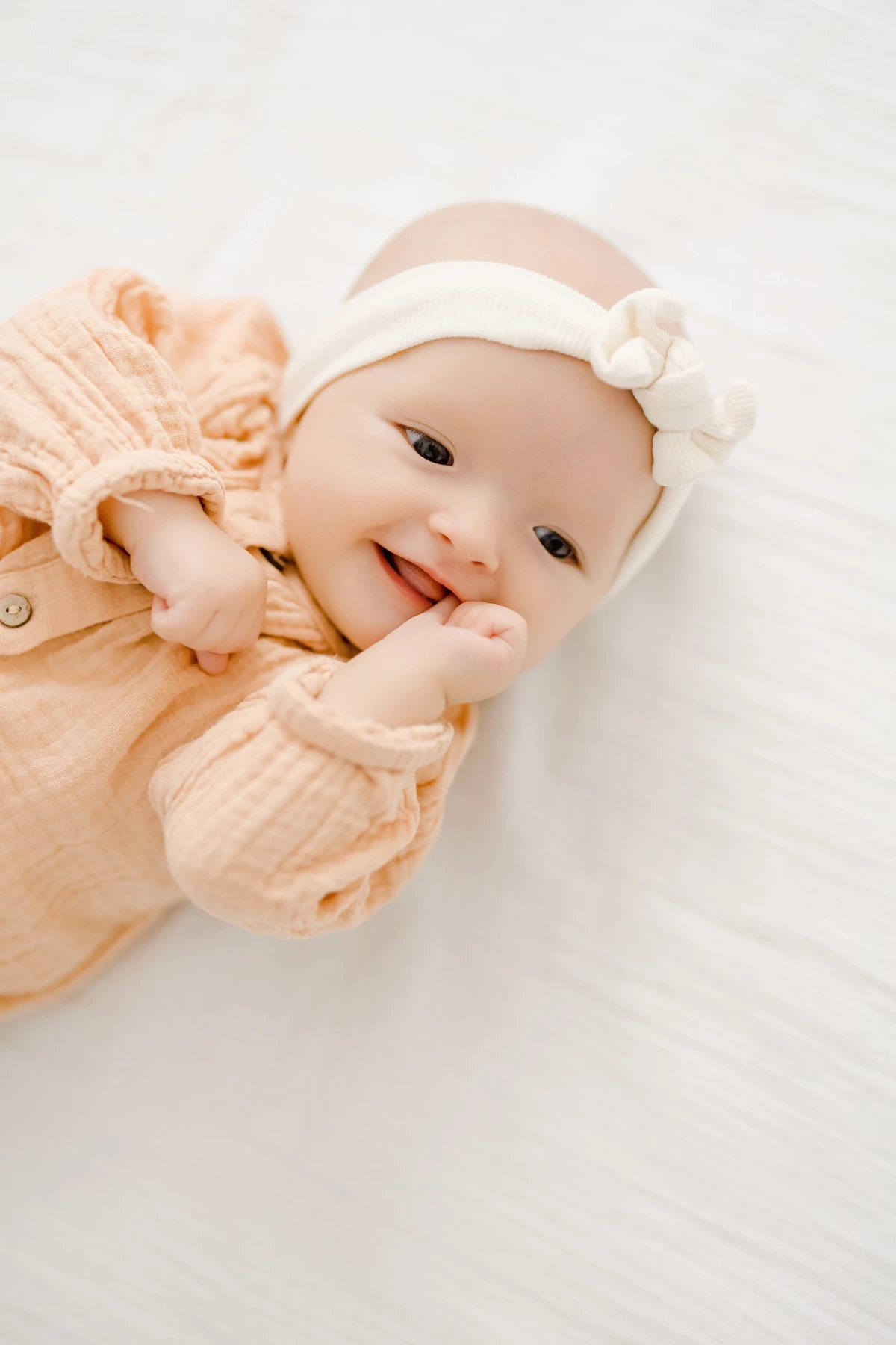 Séance naissance en studio : bébé de 4 mois sourit avec tendresse, vêtu de beige, entouré de lumière douce et d'un bandeau fleuri blanc
