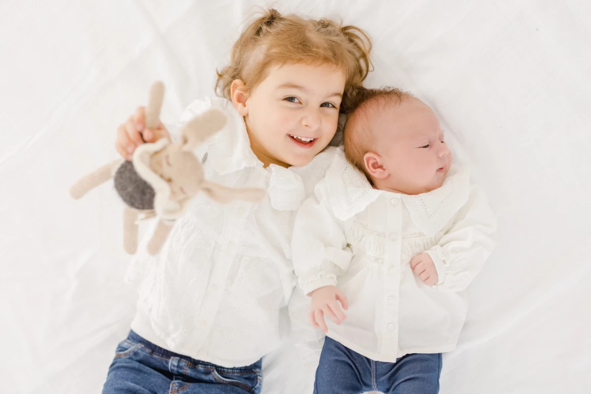 Séance naissance en studio : enfant souriante allongée près de son petit frère nouveau-né, instant de complicité fraternelle