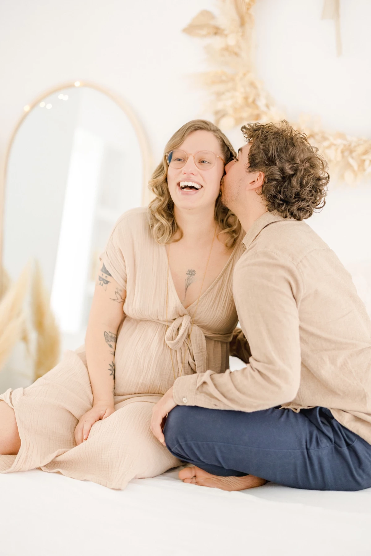 Séance grossesse en studio : couple complice partage un moment de tendresse, lui l'embrasse tendrement dans une lumière douce