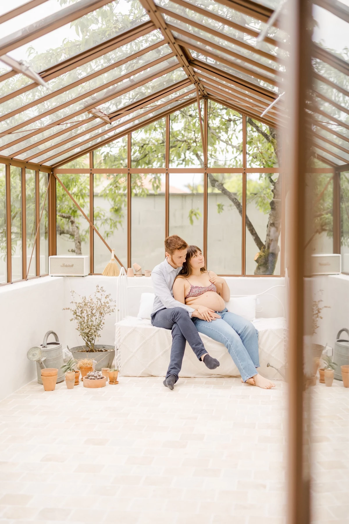 Séance grossesse sous verrière : couple enlacé partage un moment d'intimité dans un décor lumineux aux tons naturels
