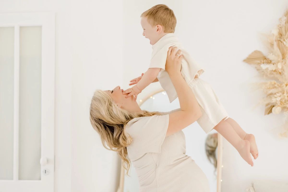 Séance famille au studio : maman soulève son jeune enfant dans les bras, instant de complicité et de joie sur fond blanc lumineux