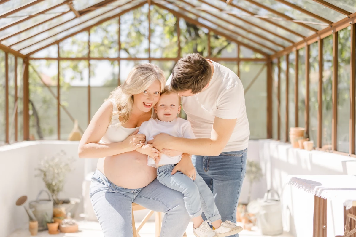 Séance grossesse en verrière : futurs parents et leur enfant partagent un moment de tendresse, lumière naturelle douce