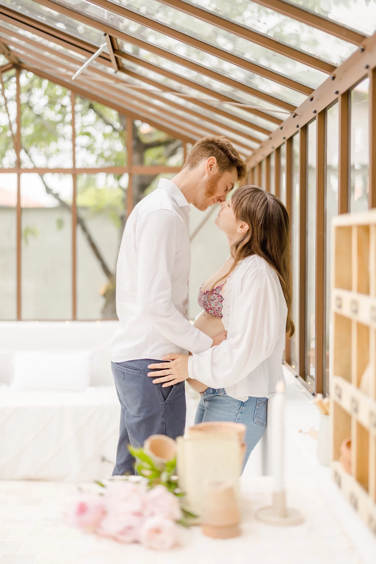 Séance grossesse en verrière : couple complice partage un moment de tendresse, lumière naturelle et douceur dans un décor champêtre