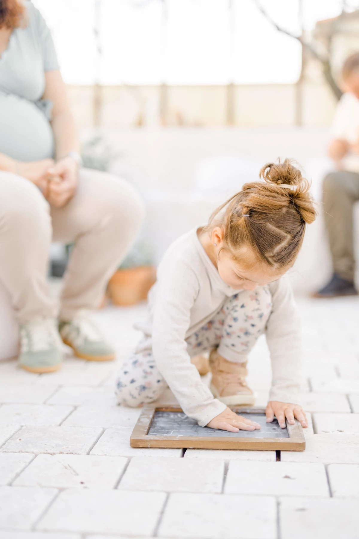 Séance grossesse famille en verrière : petite fille concentrée au premier plan, parents en arrière-plan, lumière douce naturelle