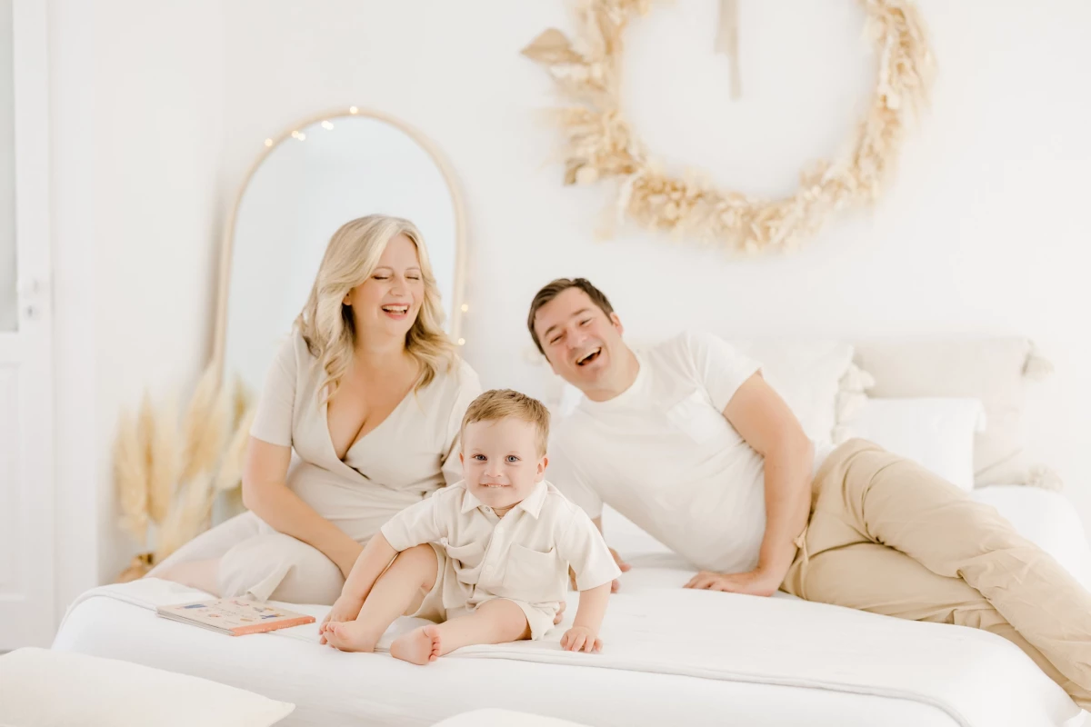 Séance grossesse au studio : famille complice sur un lit blanc, parents et enfant partagent un moment de tendresse et de joie