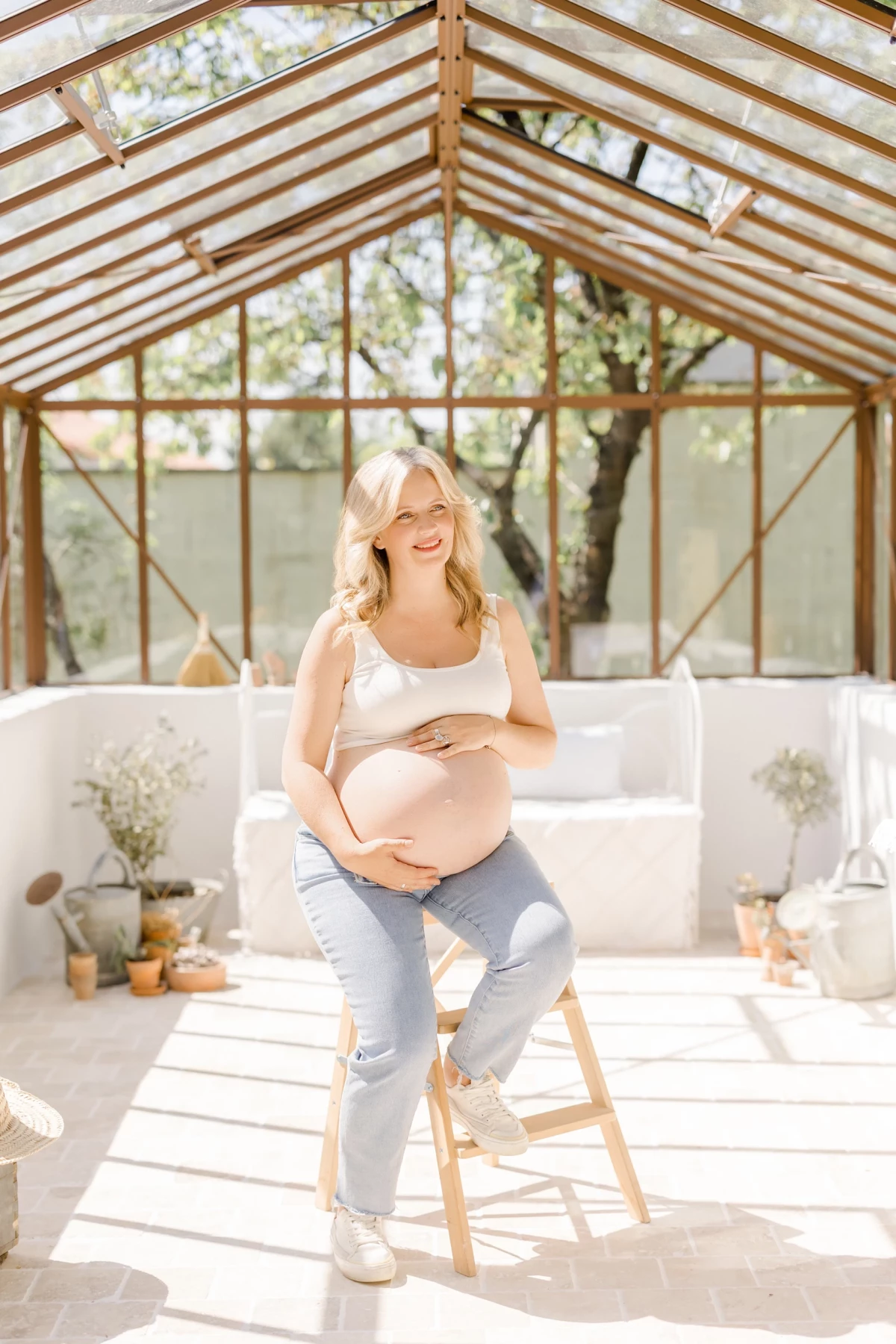 Séance grossesse au studio : future maman souriante caresse son ventre dans une verrière lumineuse, instant de tendresse et de douceur