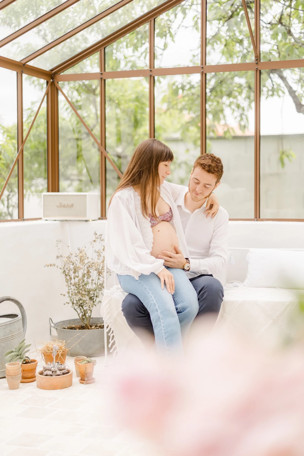 Séance grossesse en verrière : couple complice partage un moment de tendresse dans une ambiance lumineuse et végétale