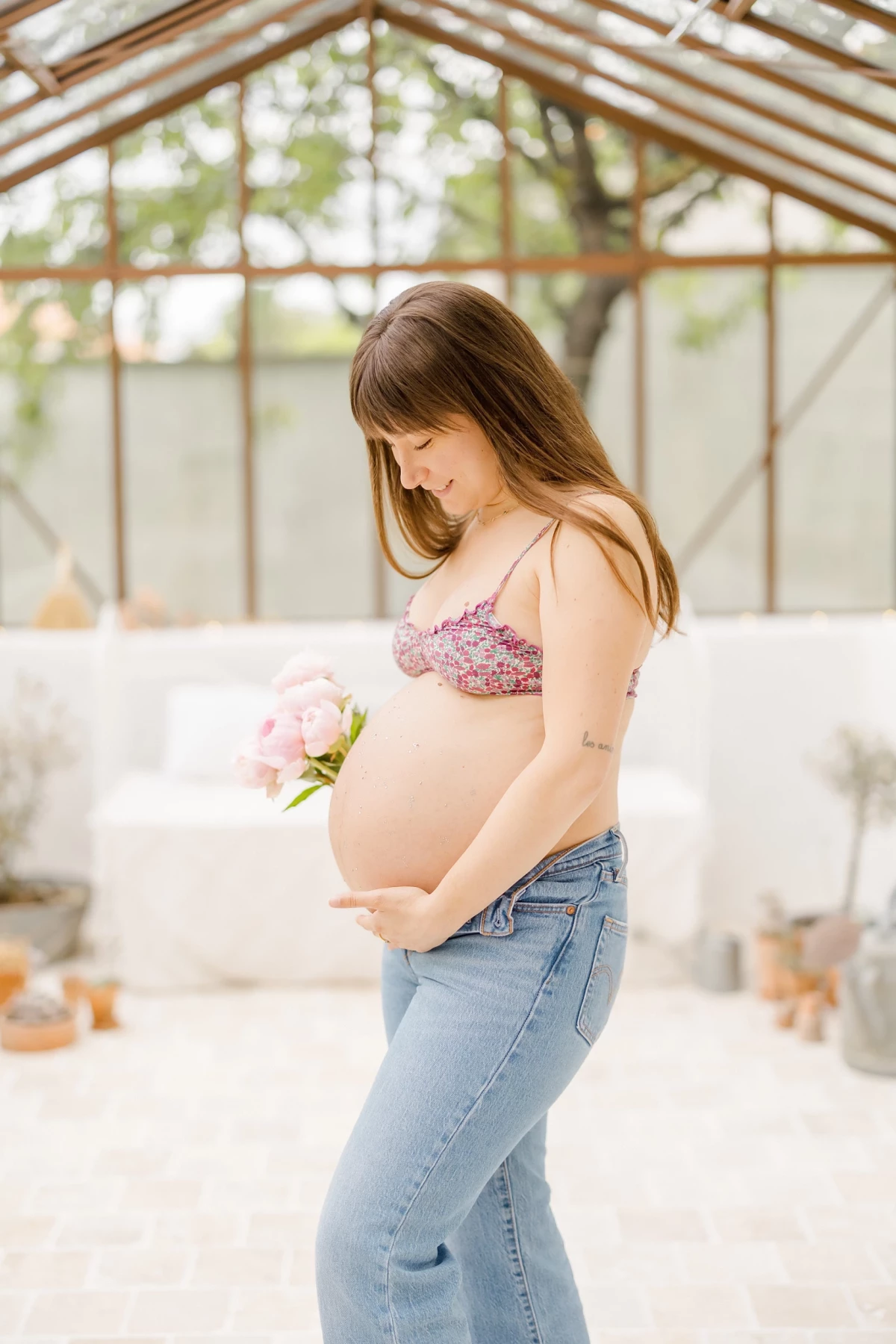 Séance grossesse en verrière : future maman en jeans et top fleuri caresse tendrement son ventre rond, tenant une pivoine rose