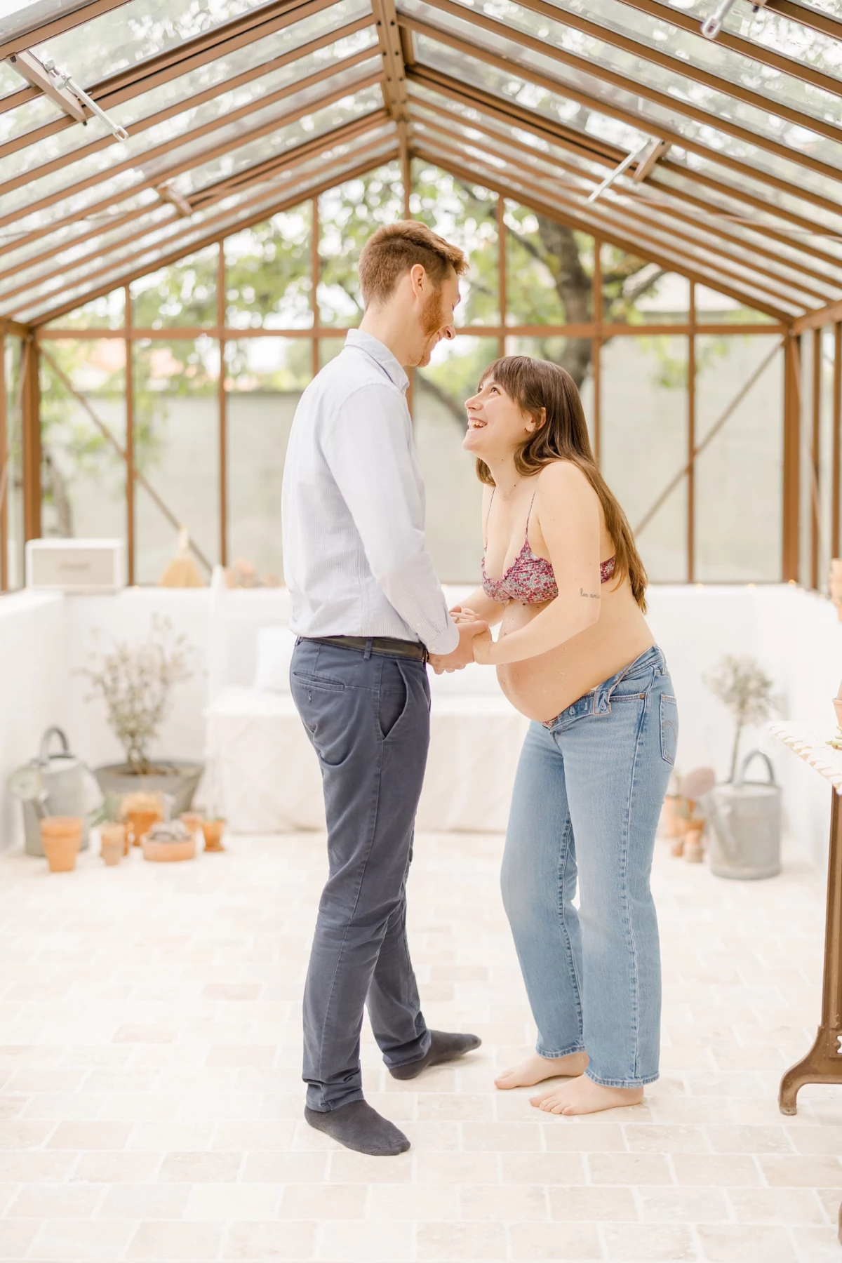 Séance grossesse au studio : couple complice partageant un moment de tendresse sous une verrière, ambiance bucolique et lumineuse