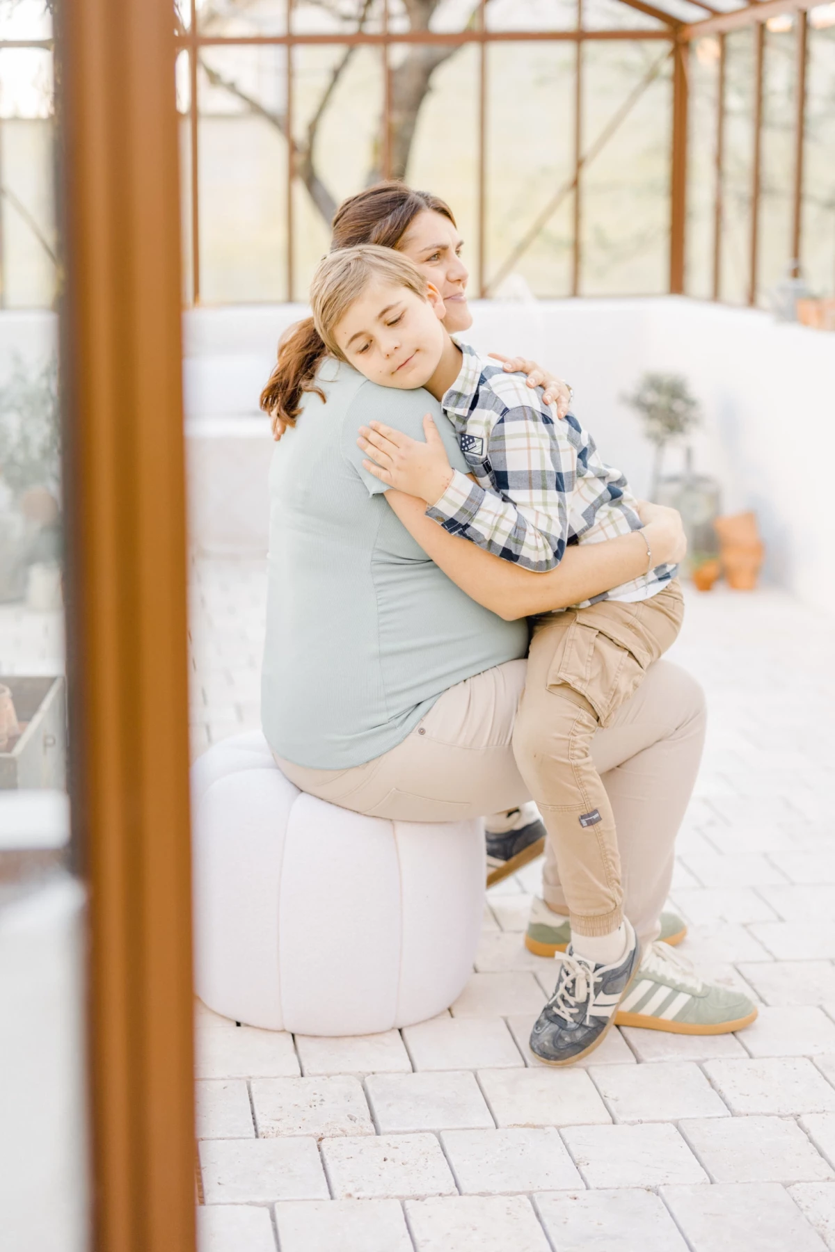 Séance grossesse en studio : enfant enlace tendrement sa maman enceinte dans une verrière lumineuse, complicité mère-fille