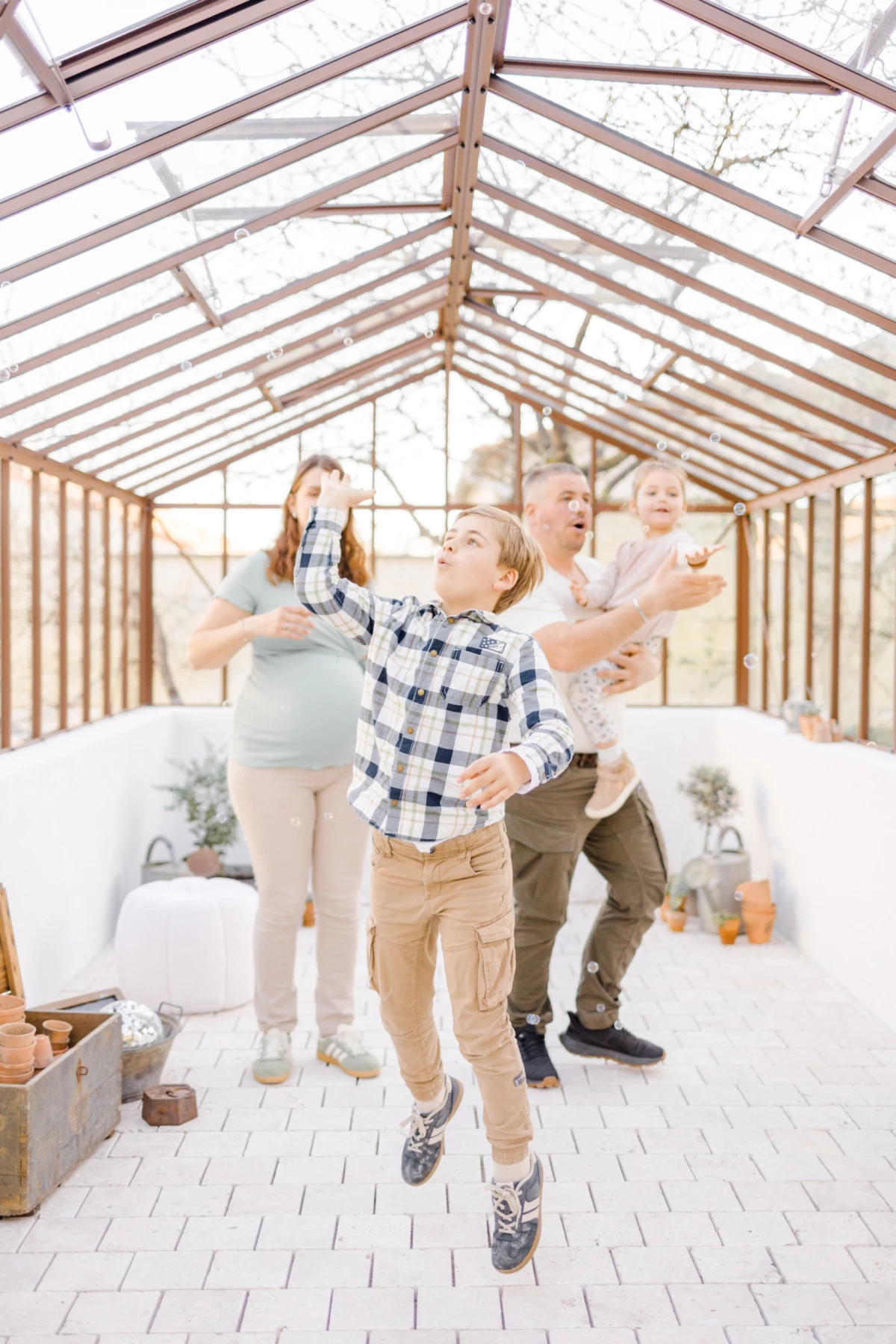 Séance famille en verrière : enfants jouent avec joie dans une serre lumineuse aux tons beige et blanc, ambiance naturelle et douce