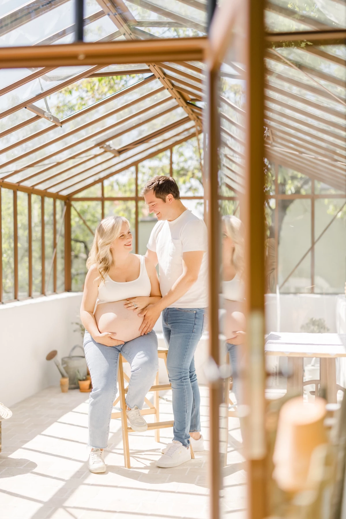 Séance grossesse au studio : couple complice sous verrière, future maman caresse son ventre, lumière douce et naturelle en Vendée