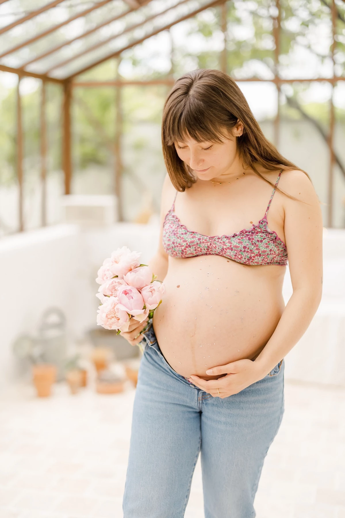 Séance grossesse en extérieur : future maman caresse son ventre rond avec tendresse, tenant un bouquet de fleurs roses sous une verrière