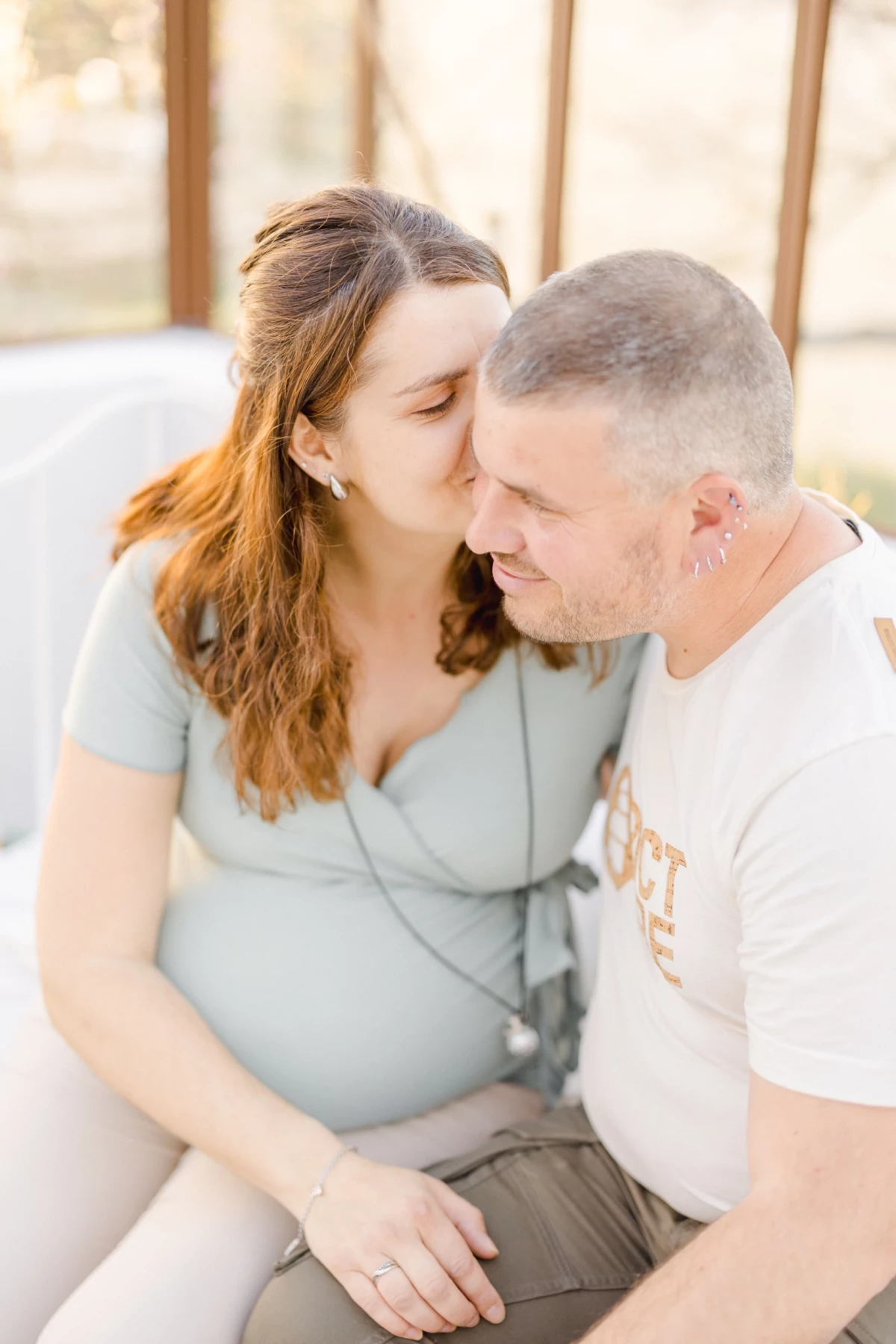 Séance grossesse en verrière : couple complice partage un moment tendre, lumière douce enveloppant la future maman en robe grise