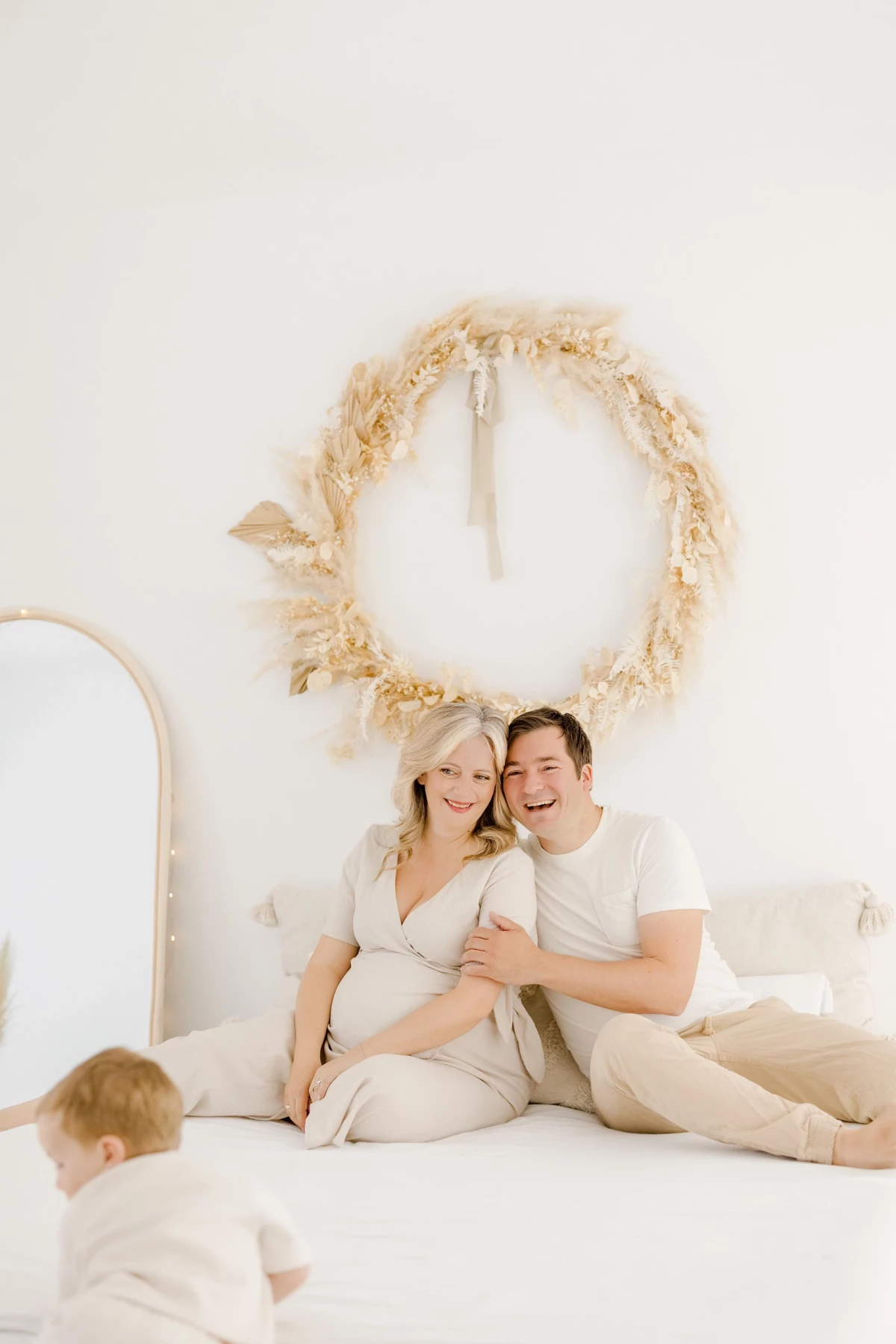 Séance grossesse studio : couple complice et leur enfant sur un lit blanc sous une couronne de fleurs séchées, ambiance douce et beige