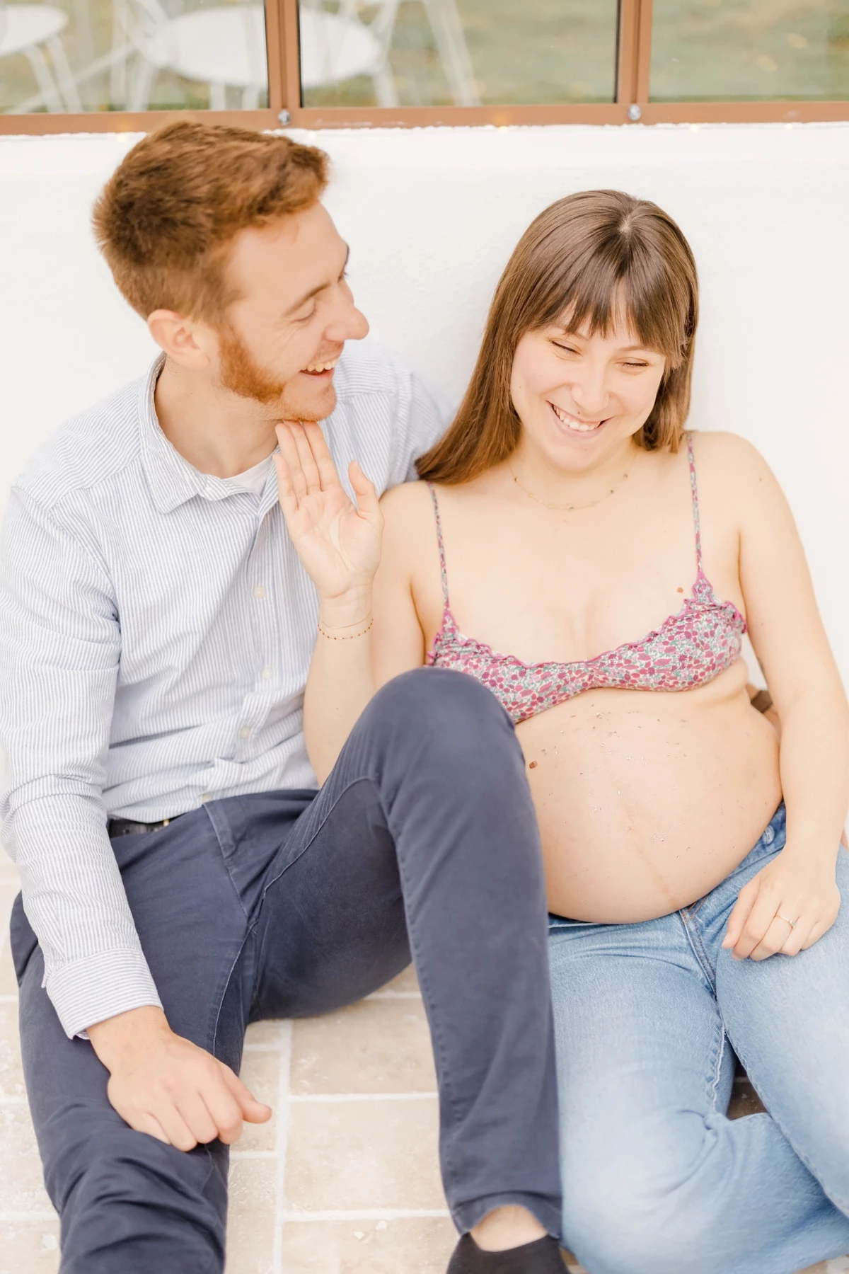 Séance grossesse extérieure : couple complice sous verrière, future maman dévoile son ventre rond dans une lumière douce et naturelle