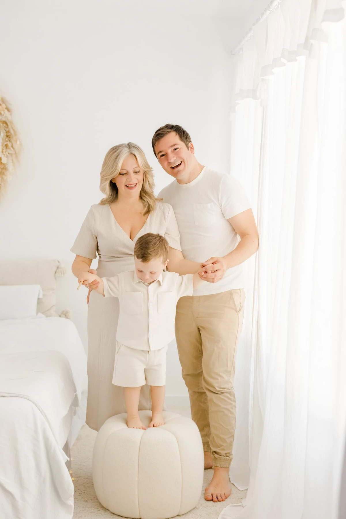 Séance grossesse en studio : famille complice, enfant debout entre ses parents, ambiance douce et lumineuse en tons beige et blanc