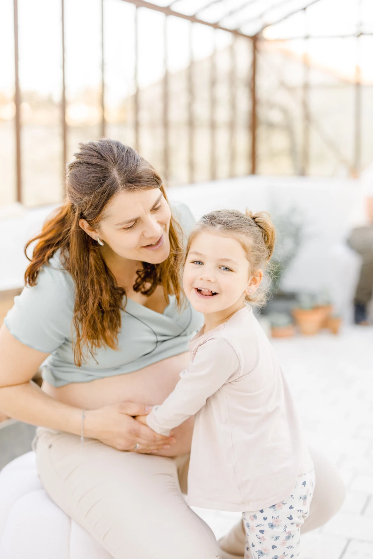 Séance grossesse famille : maman enceinte et sa fille partagent un câlin complice dans une verrière lumineuse en Vendée