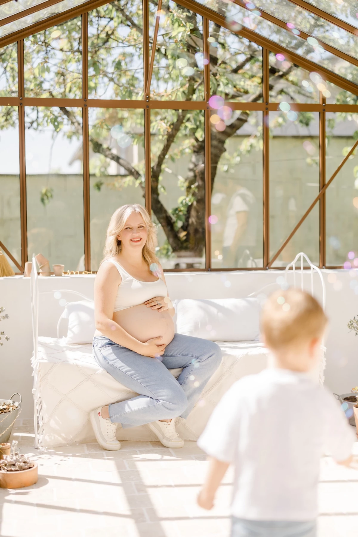 Séance grossesse en studio : future maman souriante observe son enfant jouer dans une verrière lumineuse, moment de tendresse familiale