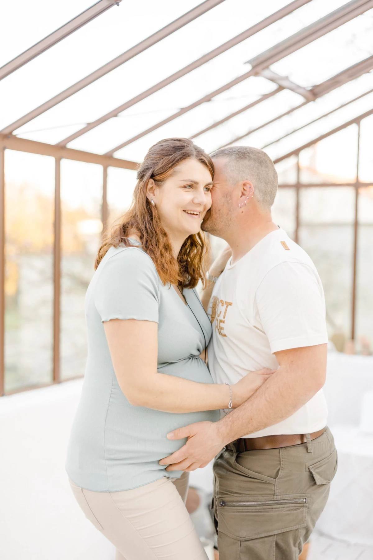 Séance grossesse en verrière : couple complice partage un moment de tendresse, baigné de lumière naturelle douce