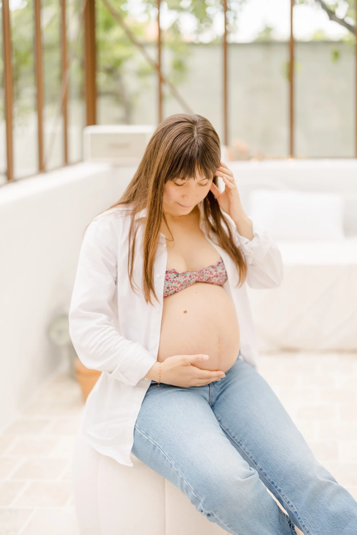 Séance maternité en extérieur : future maman caresse son ventre rond avec tendresse sous une verrière lumineuse en Vendée