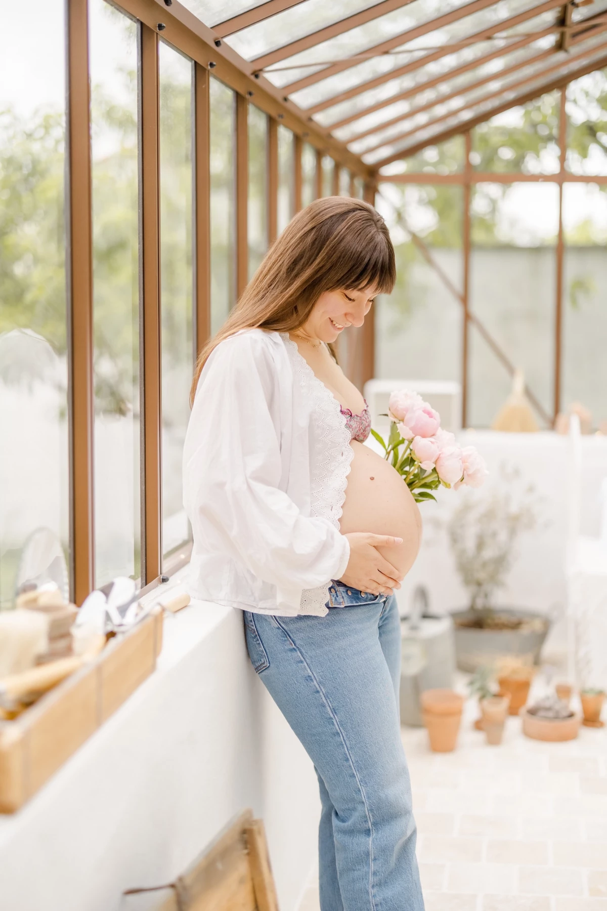 Séance grossesse en verrière : future maman enlace son ventre rond avec un bouquet de pivoines roses, lumière douce et naturelle