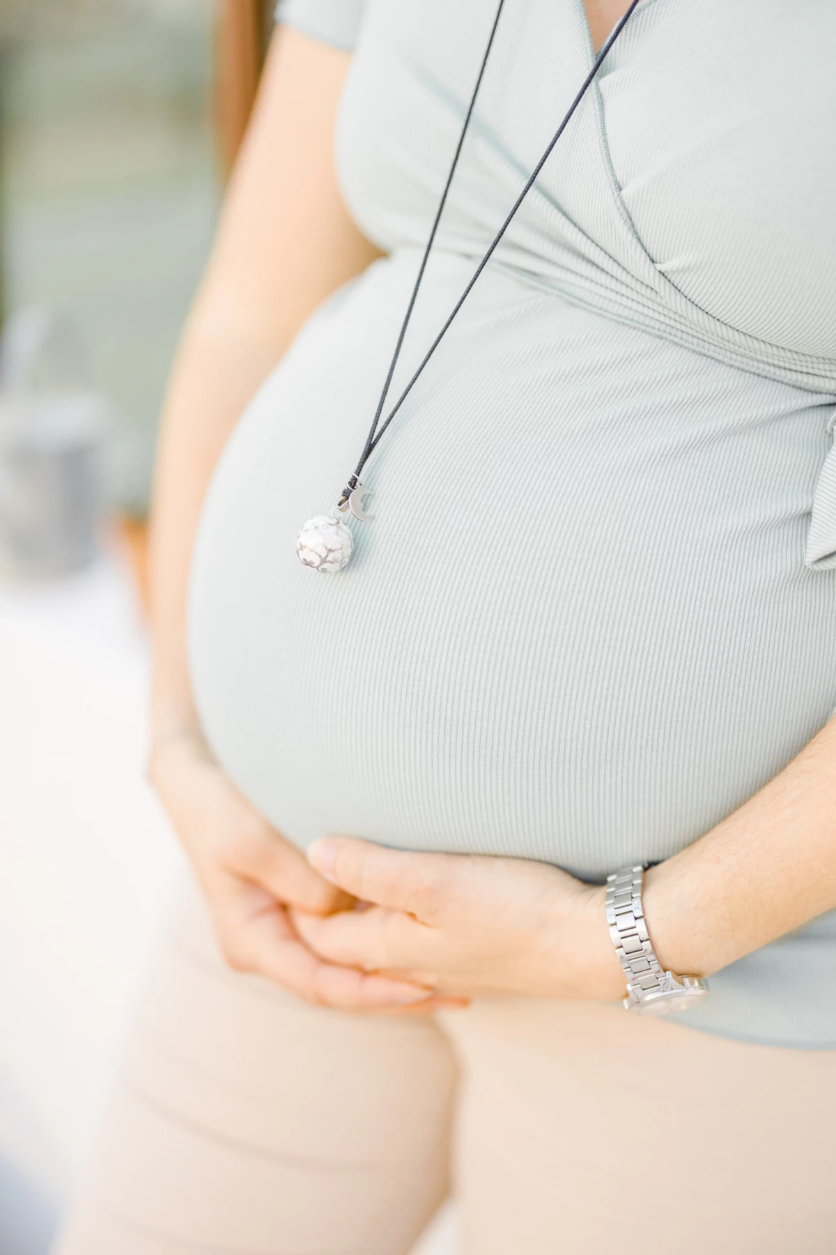 Séance grossesse en studio : future maman caresse son ventre rond avec tendresse, portant un collier délicat sur une tenue grise