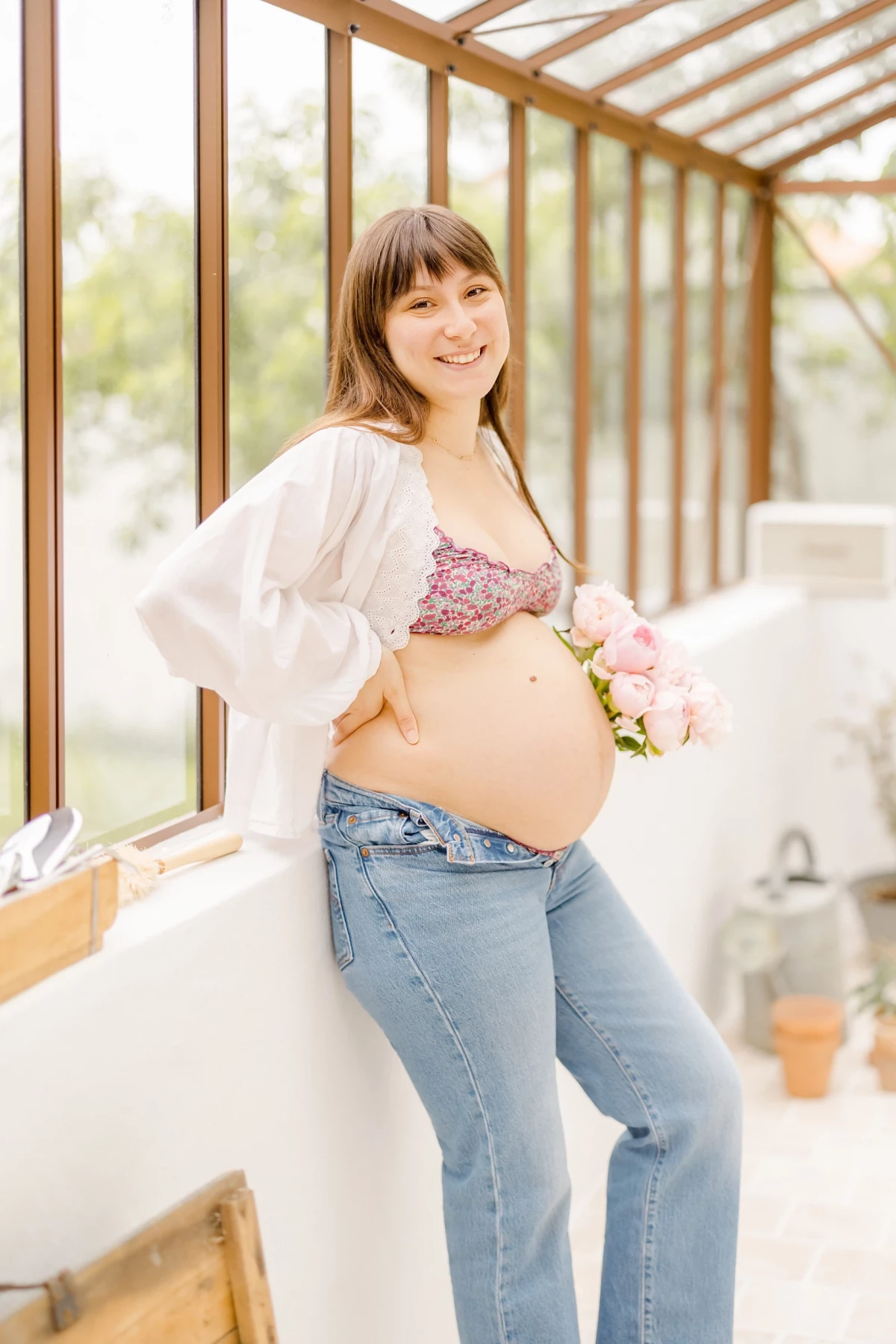 Séance grossesse en verrière : future maman souriante caresse son ventre rond, bouquet de fleurs à la main, lumière douce printanière