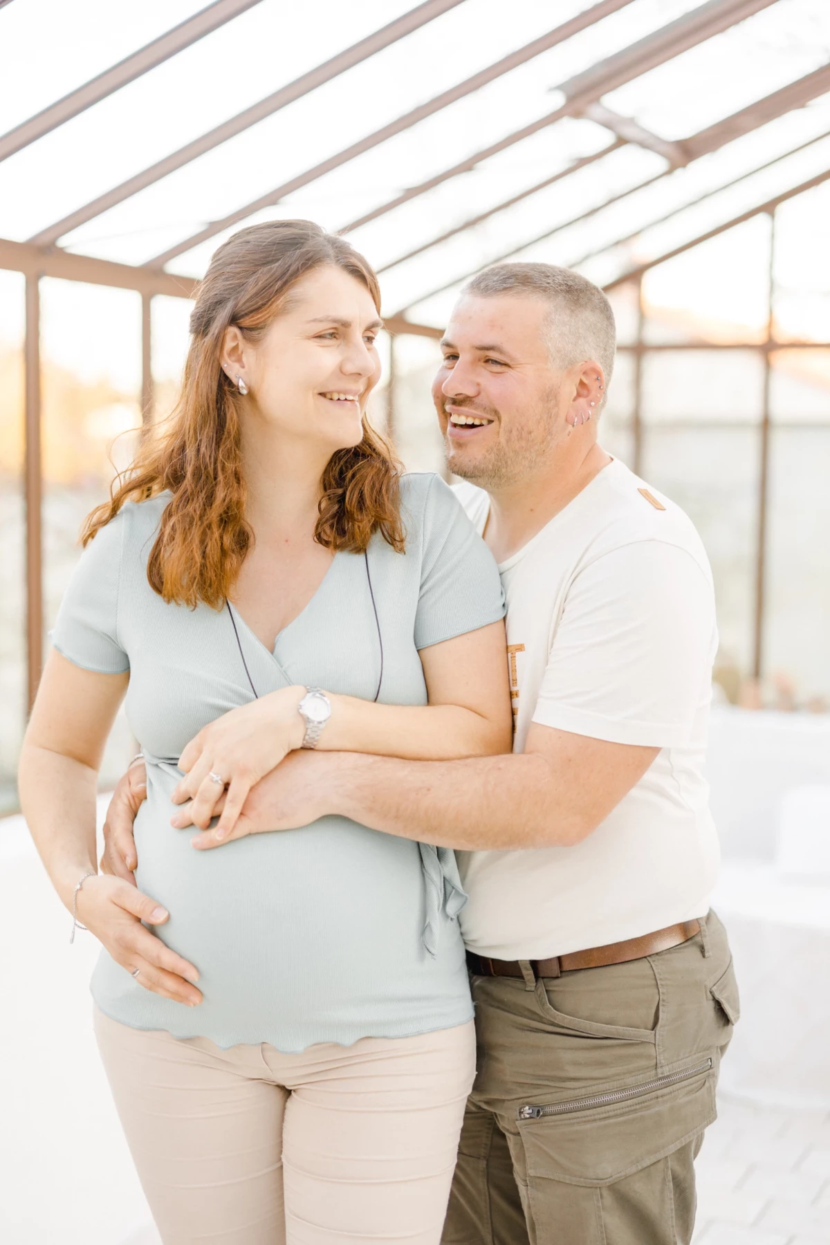 Séance grossesse en extérieur : couple complice sourit dans une verrière lumineuse, ambiance douce et naturelle en Vendée