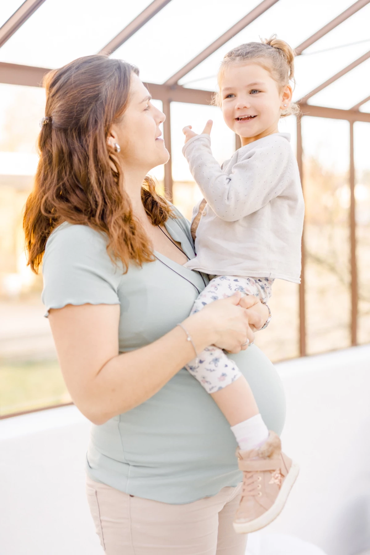 Séance grossesse famille en extérieur : maman enceinte porte sa fille avec tendresse sous une verrière lumineuse en Vendée