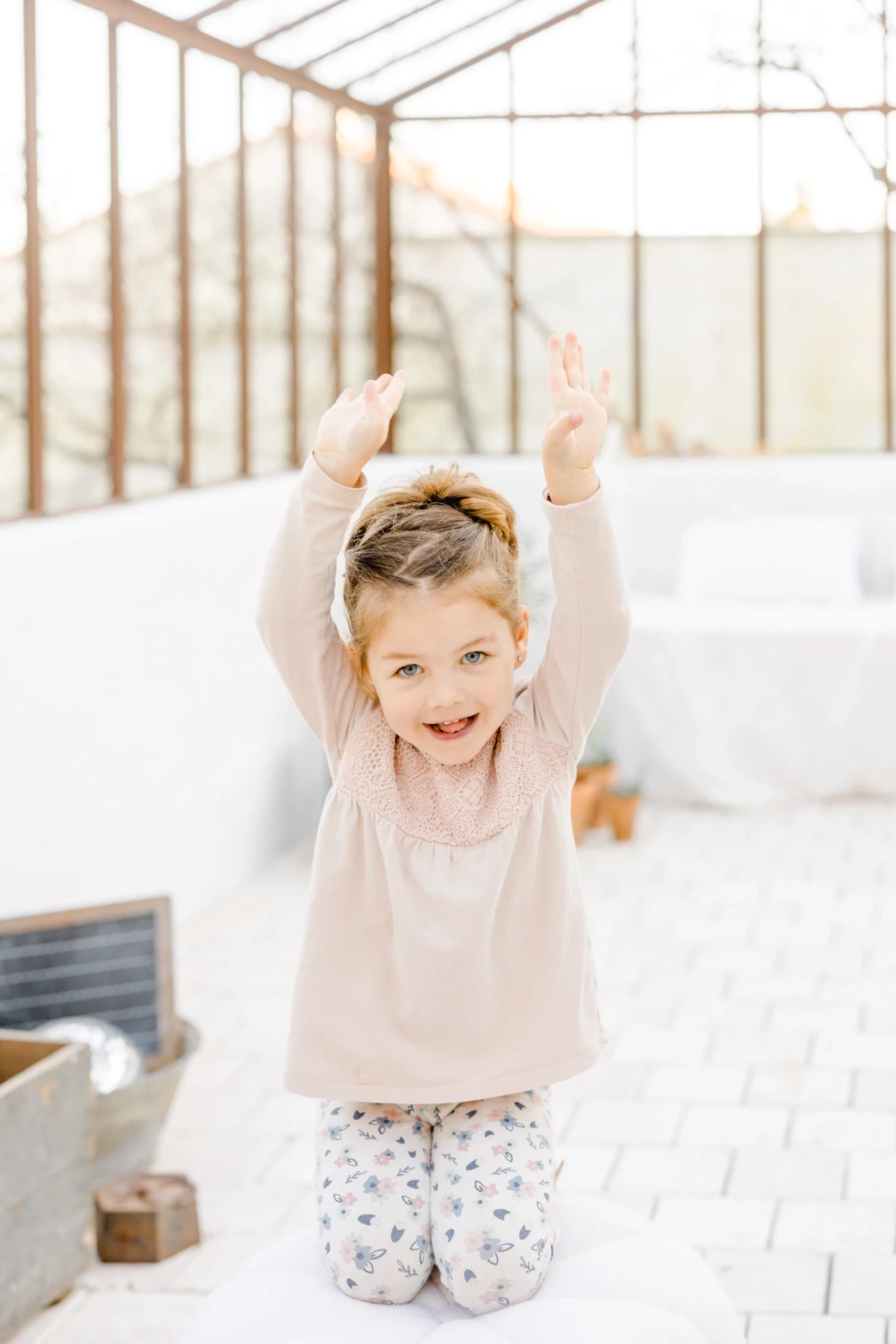 Séance famille en studio : petite fille joyeuse les bras levés dans une verrière lumineuse, instant de bonheur enfantin