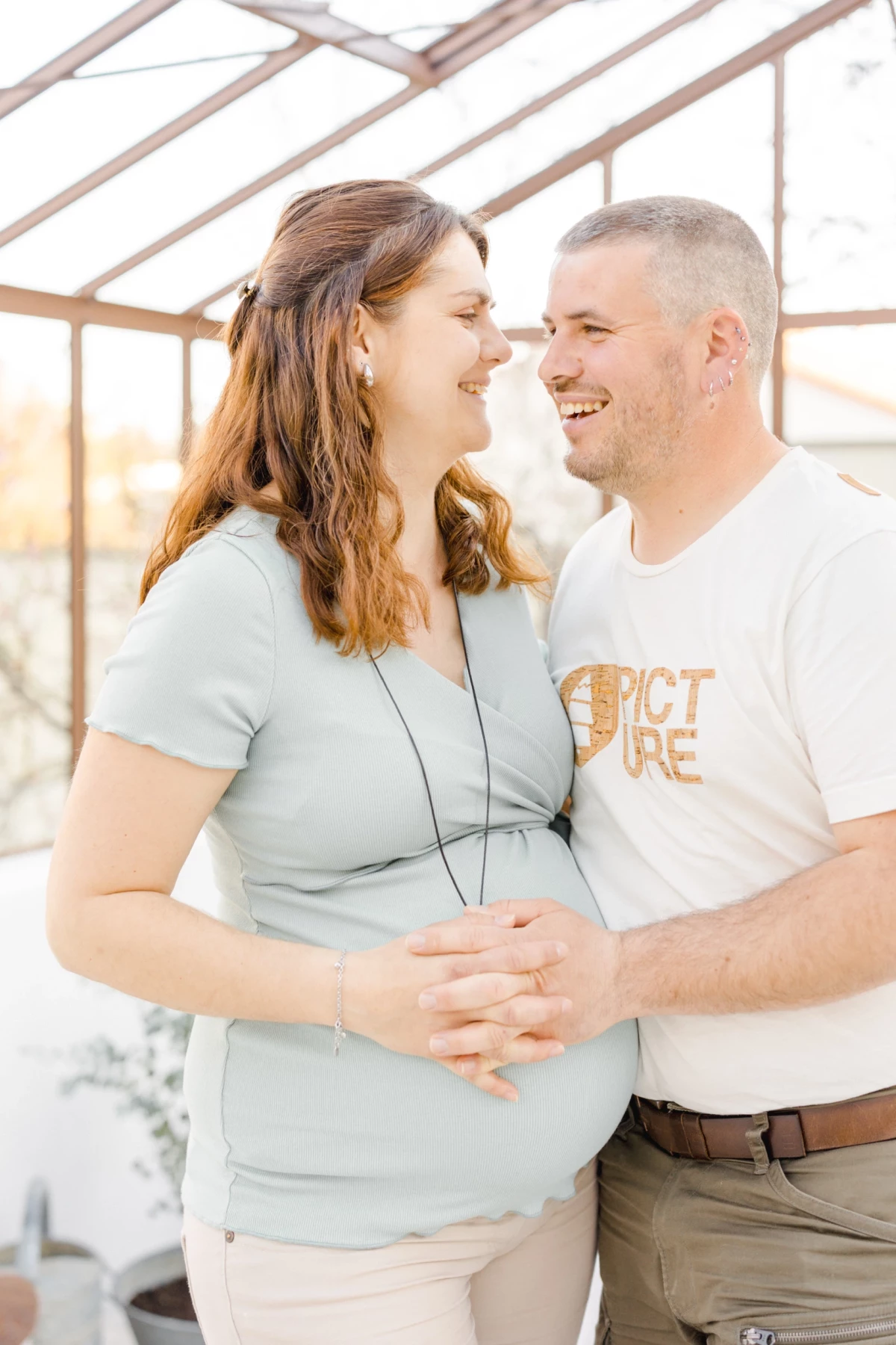 Séance grossesse en Vendée : couple complice partage un moment de tendresse dans une verrière lumineuse, mains sur le ventre rond