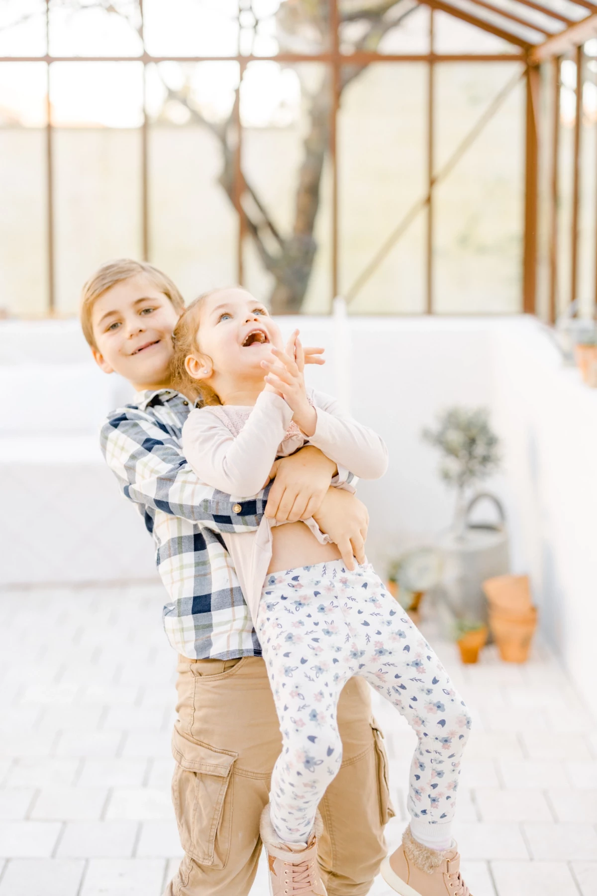 Séance famille au studio : frère et sœur rient ensemble dans une verrière lumineuse, moment de complicité spontanée et joyeuse