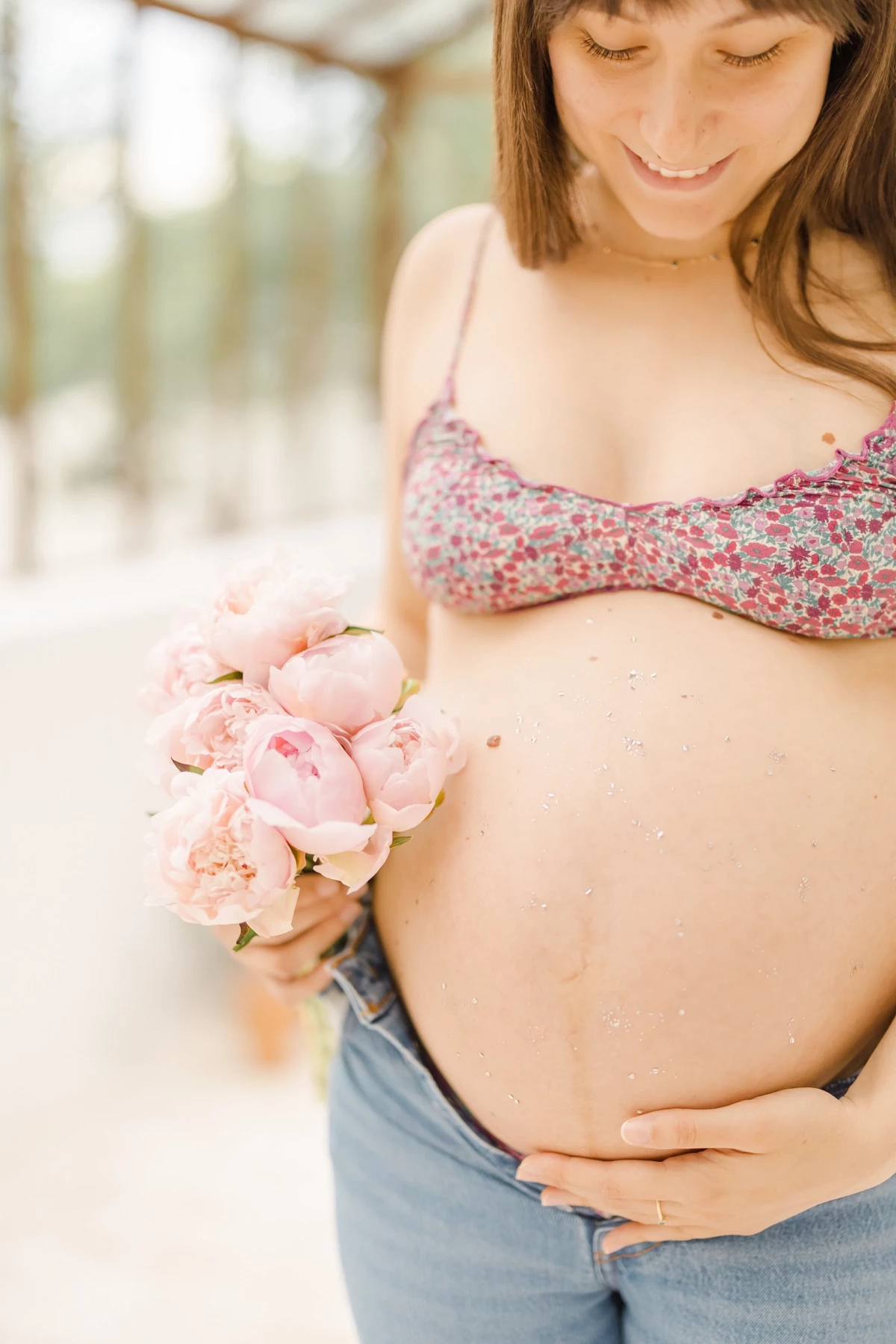 Séance grossesse extérieur : future maman souriante tenant un bouquet de pivoines roses, ambiance printanière et bucolique