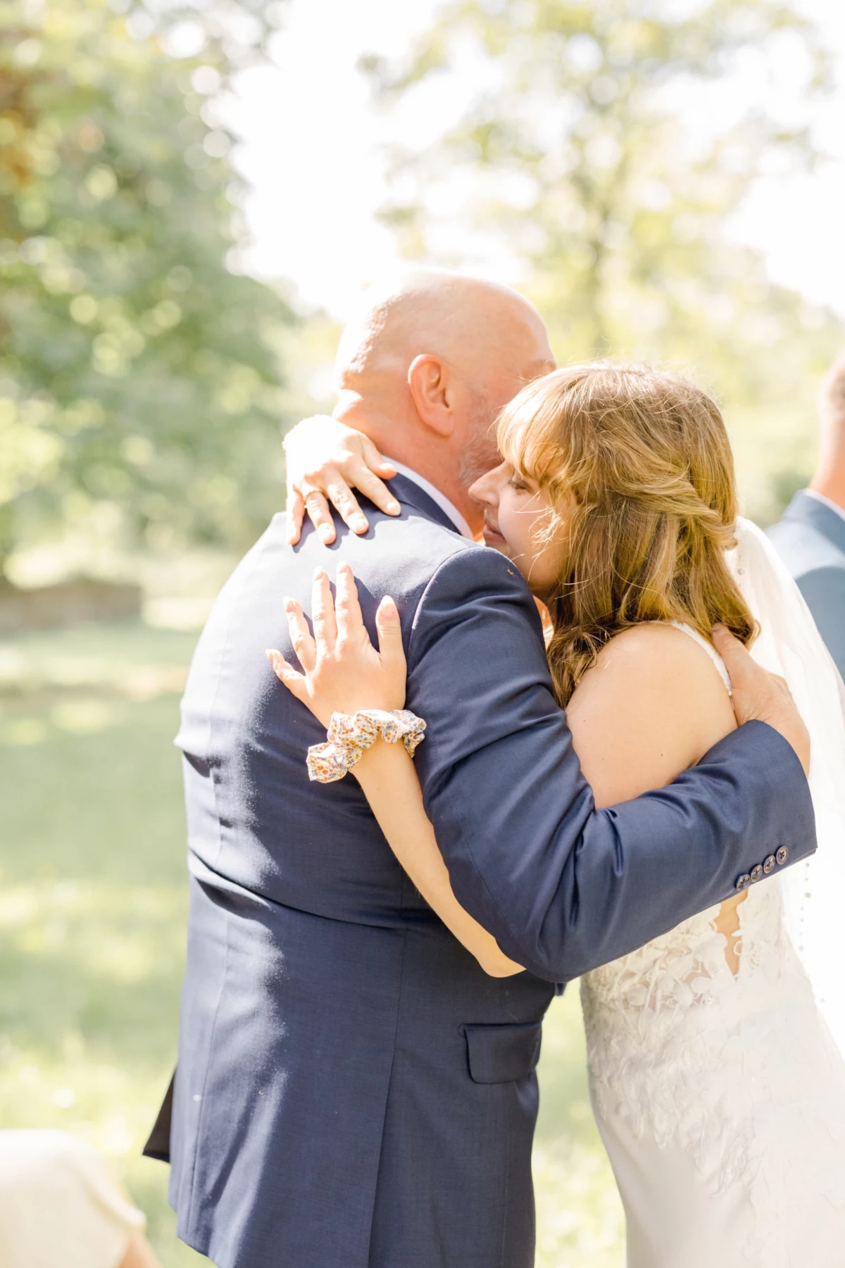 Mariée enlace tendrement son père en costume bleu marine lors de la cérémonie de mariage en extérieur