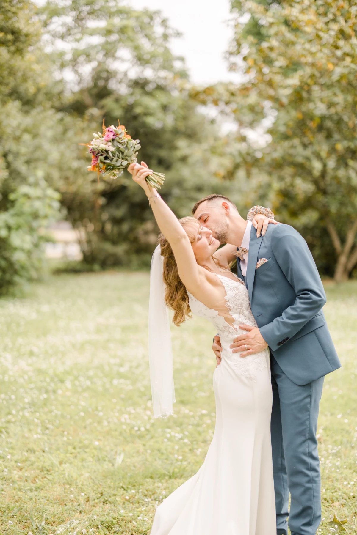 Mariée levant son bouquet au ciel pendant que son époux l'embrasse tendrement dans un parc verdoyant