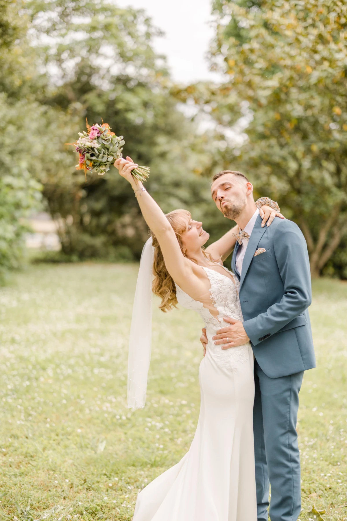 Couple de mariés célébrant dans un jardin verdoyant, la mariée brandissant son bouquet vers le ciel