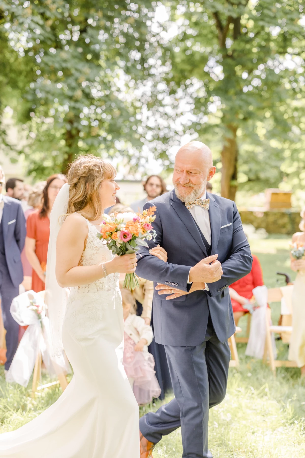 Mariée en robe blanche au bras de son père en costume bleu marine, remontant l'allée avec un bouquet coloré