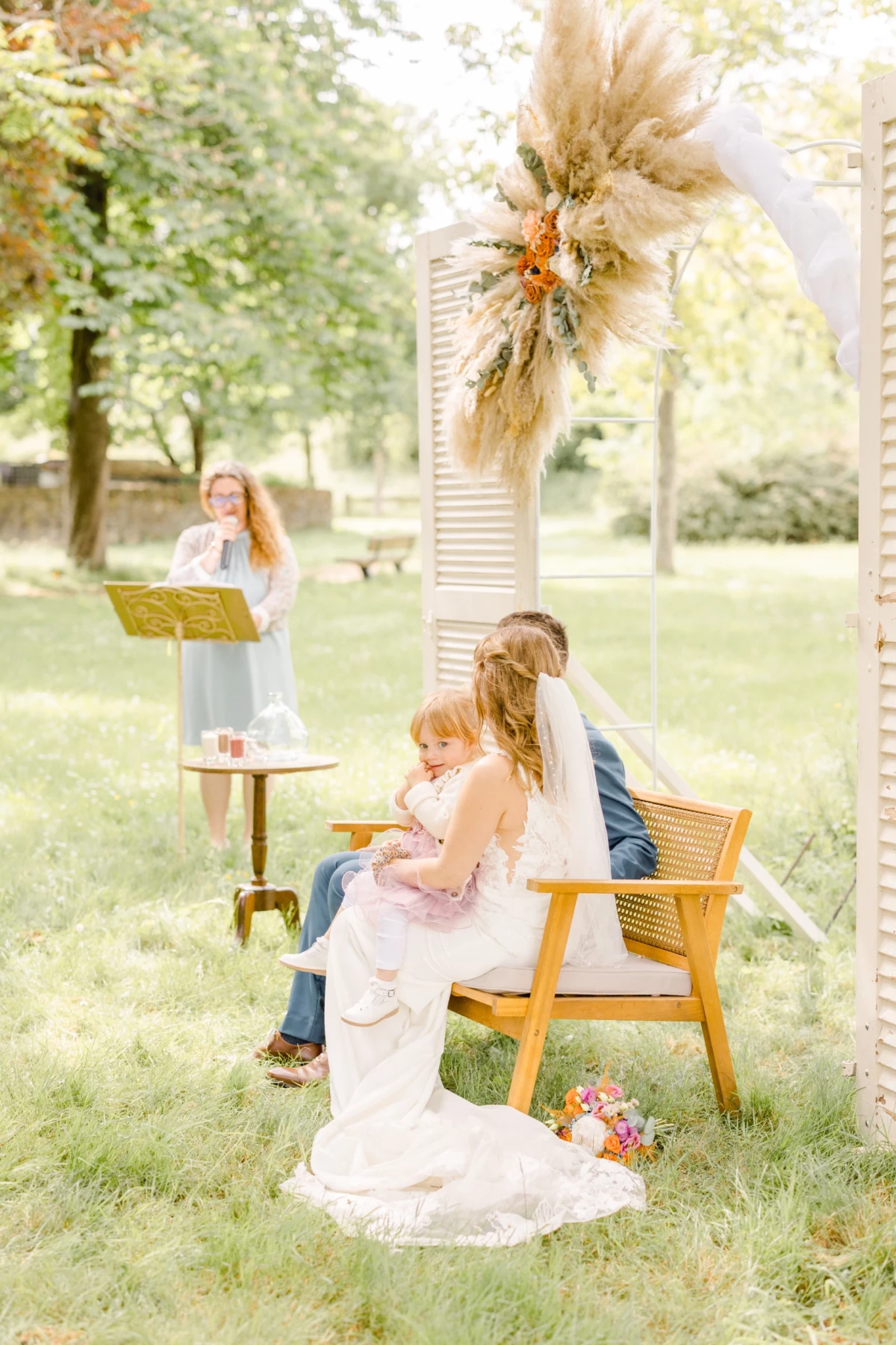 Cérémonie laïque en plein air avec couple marié assis devant une arche fleurie et officiant lisant un texte