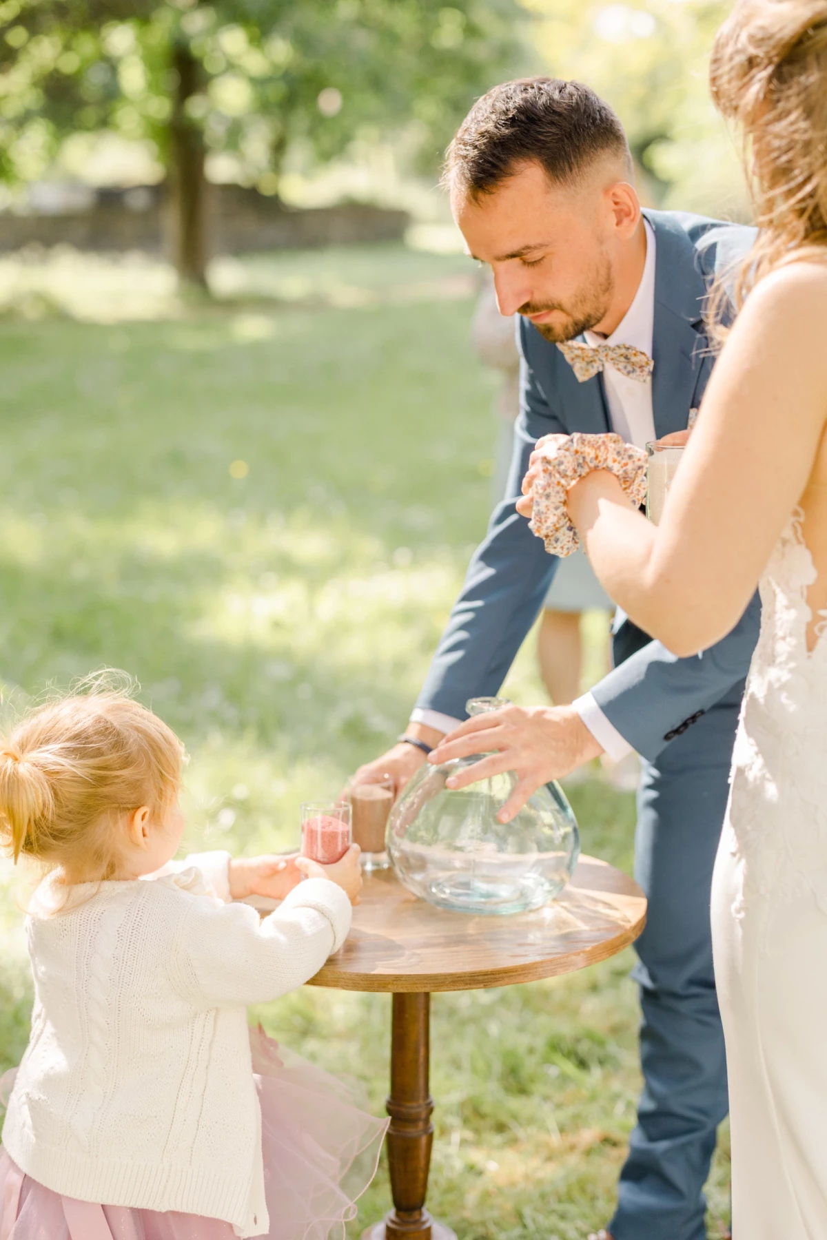 Couple de mariés et enfant versant du sable coloré dans un vase lors d'une cérémonie laïque en extérieur