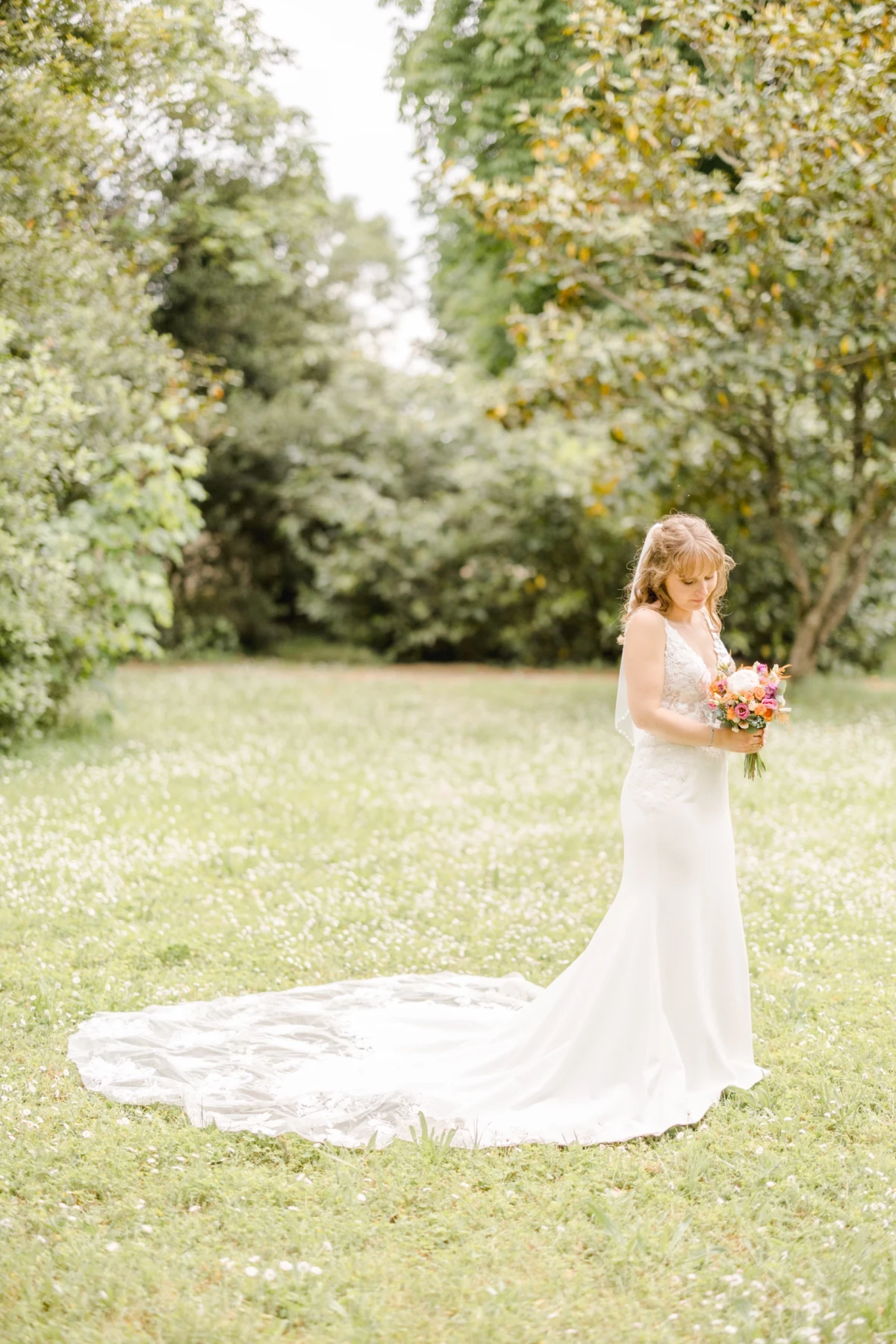 Mariée en robe blanche à longue traîne tenant un bouquet coloré dans un jardin verdoyant