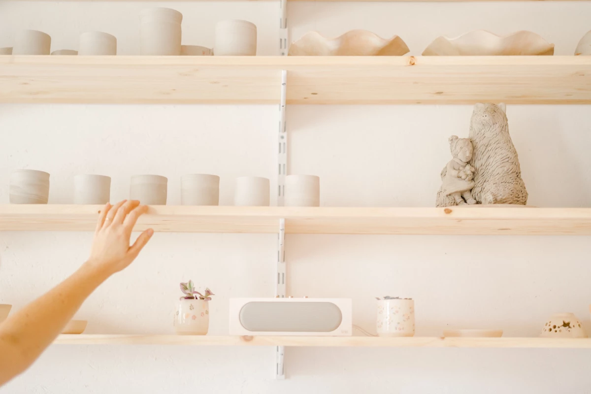 Portrait corporate dans un atelier de céramiste : main tendue vers des créations artisanales disposées sur des étagères en bois naturel