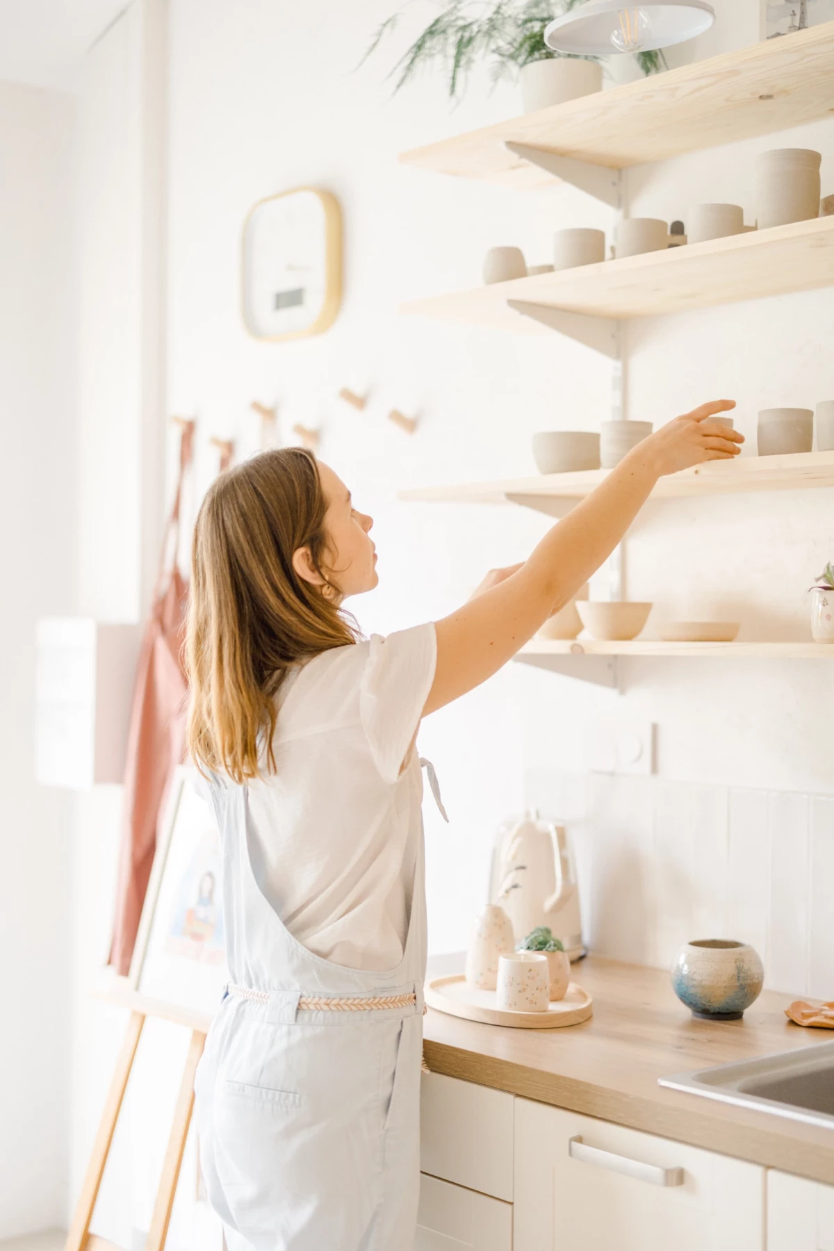 Portrait corporate dans atelier céramique : artiste arrange ses créations sur étagères en bois, lumière douce et naturelle en Vendée