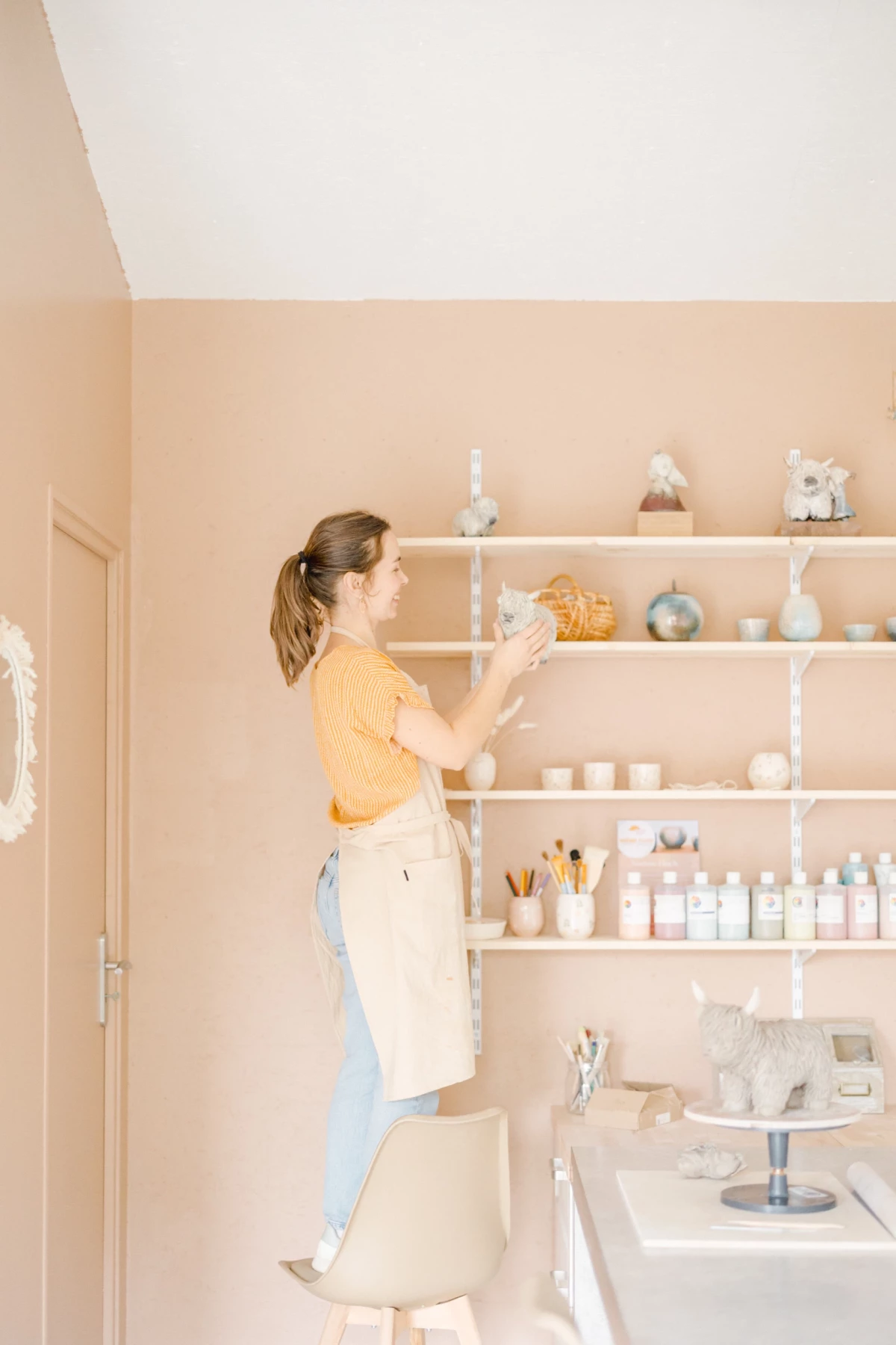 Portrait professionnel en studio : artiste céramiste et aquarelliste arrange ses créations sur des étagères dans une ambiance lumineuse et épurée