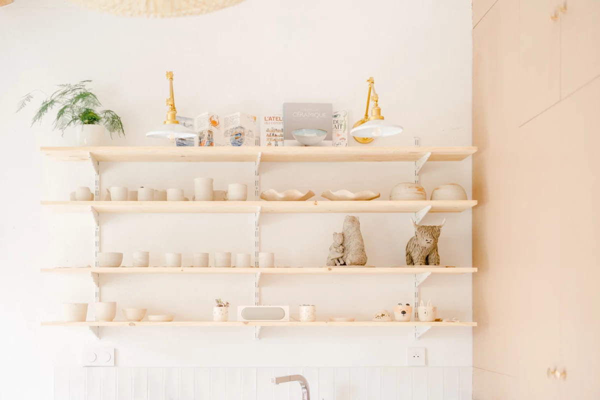 Portrait corporate dans un atelier céramiste : étagères blanches exposant céramiques artisanales et aquarelles, lumière douce naturelle