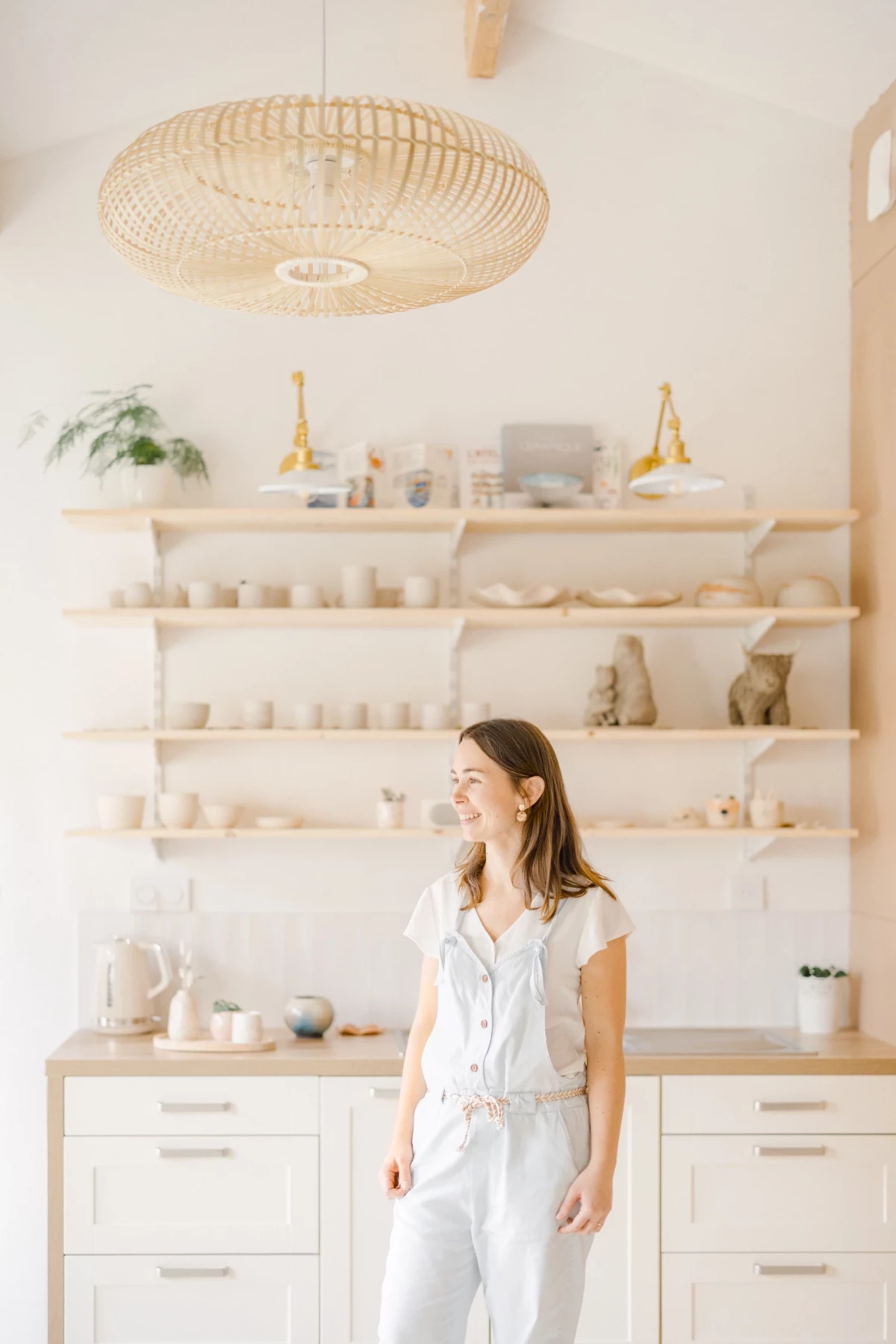 Portrait professionnel d'une céramiste dans son atelier lumineux, entourée de créations artisanales sur étagères blanches