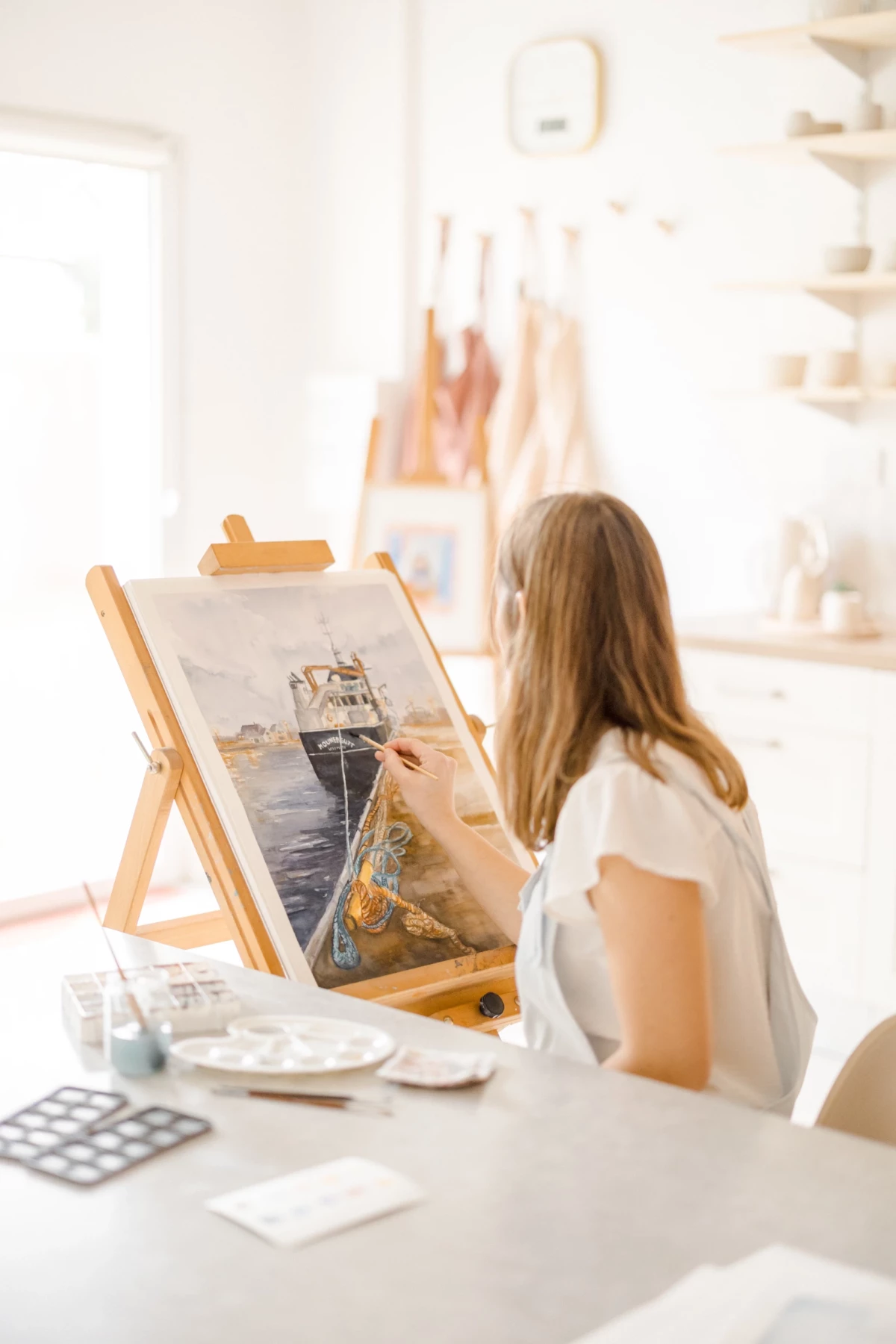 Portrait corporate en atelier : artiste céramiste et aquarelliste peint une œuvre maritime dans son espace lumineux et chaleureux